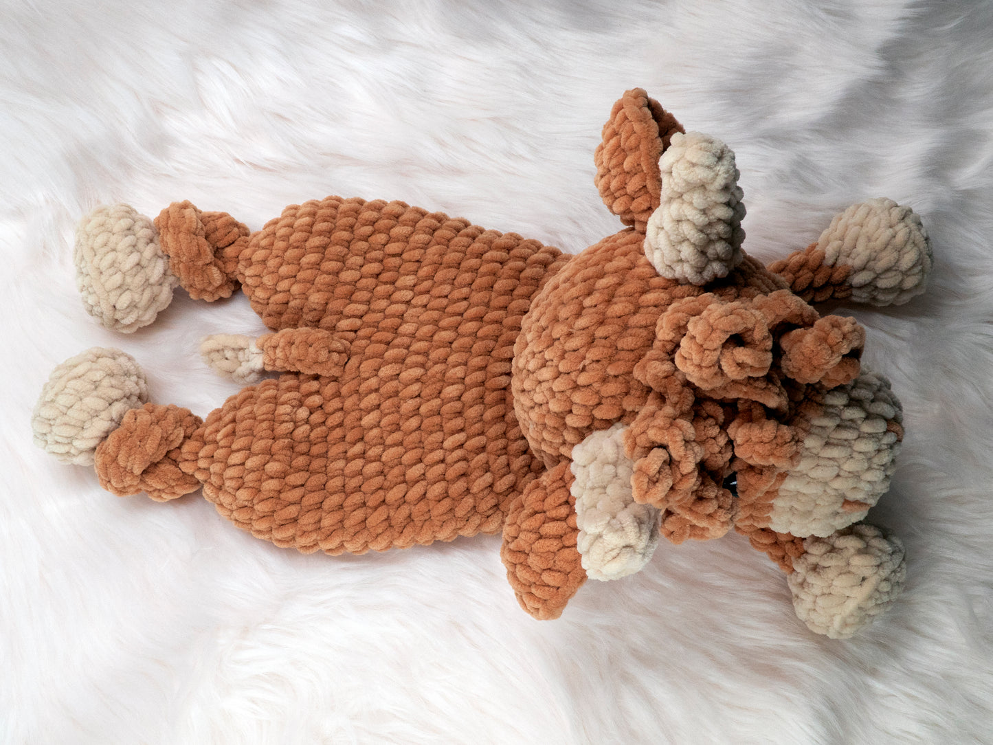 a top down view of crochet highland cow lovey made of chenille yarn laying down on its stomach on a white fur carpet.