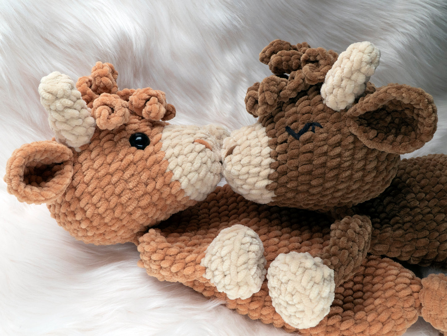 Two crochet highland cow loveys made of chenille yarn touching noses as if kissing. One shows the optional embroidered eye instructions available in the crochet pattern.