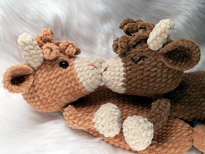 Two crochet highland cow loveys made of chenille yarn touching noses as if kissing. One shows the optional embroidered eye instructions available in the crochet pattern.