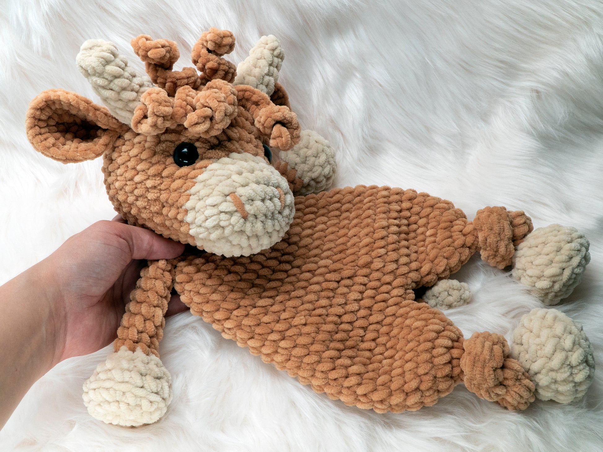 a crochet highland cow lovey made of chenille yarn being picked up by hand that is coming into view from the left hand side of the image, to show a size comparision.
