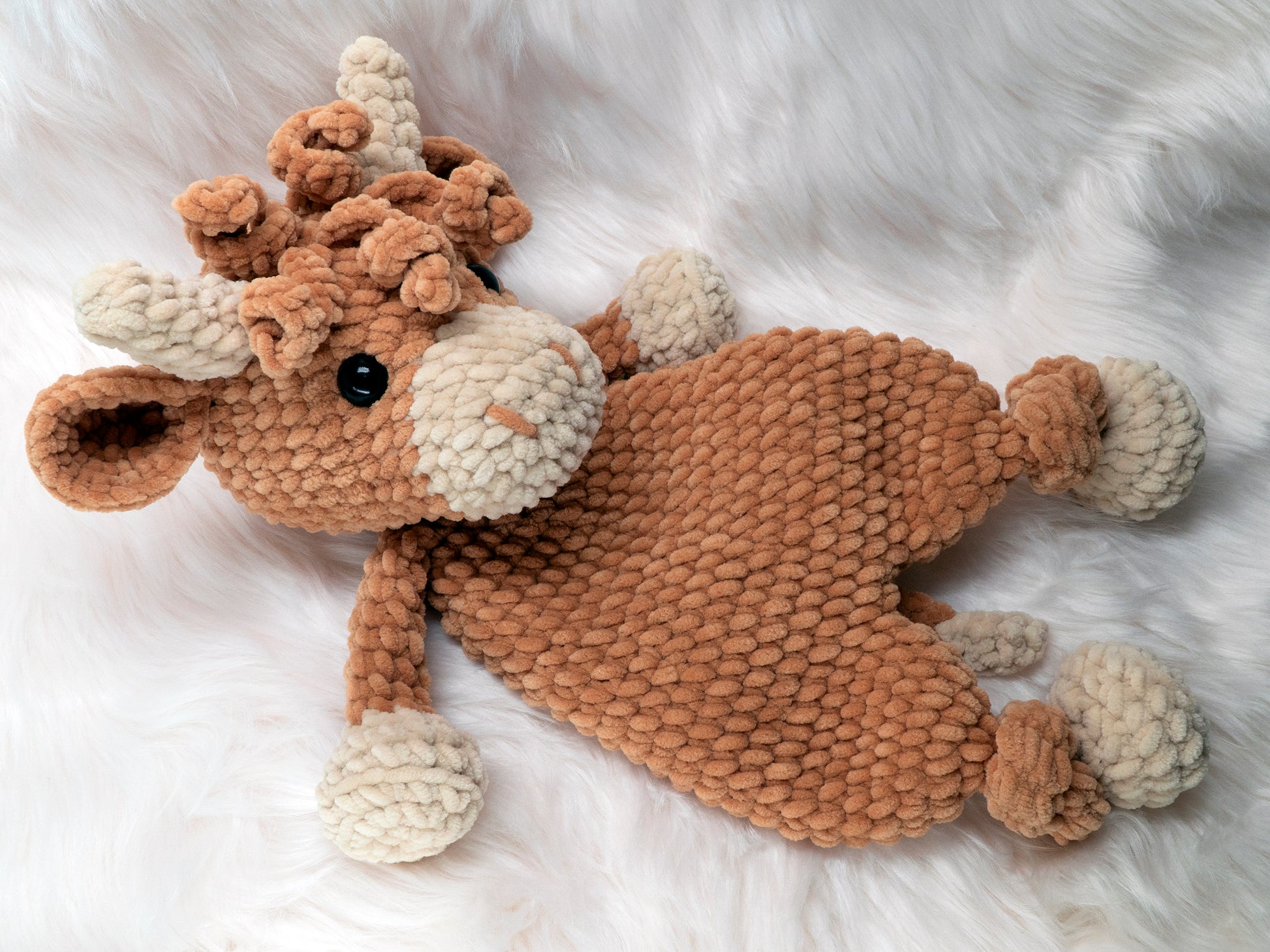 a crochet highland cow lovey made of chenille yarn laying down on a white fur background staring up at the viewer.