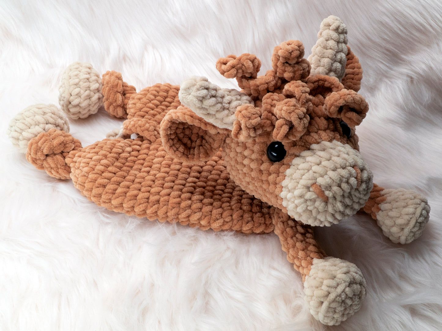 a crochet highland cow lovey made of chenille yarn laying down on its stomach on a white fur carpet staring up at the viewer.